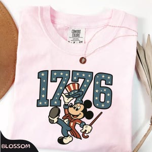 May include: A light pink t-shirt featuring the number "1776" in blue with white stars. A cartoon character in patriotic attire is below the numbers. A Comfort Colors tag is visible at the neckline.
