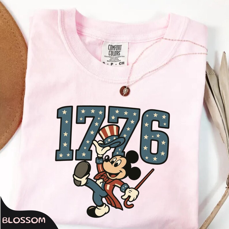 May include: A light pink t-shirt featuring the number "1776" in blue with white stars. A cartoon character in patriotic attire is below the numbers. A Comfort Colors tag is visible at the neckline.