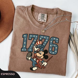 May include: Espresso-colored t-shirt featuring the number "1776" in blue with white stars, and a cartoon character in patriotic attire. The shirt has a crew neck and short sleeves. A silver necklace with a pendant is draped on the shirt.