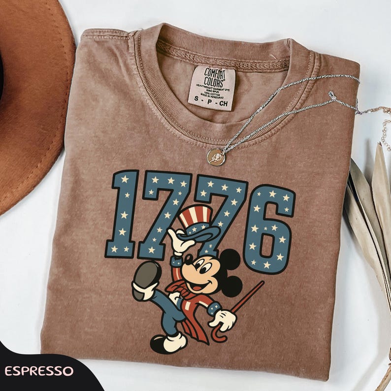 May include: Espresso-colored t-shirt featuring the number "1776" in blue with white stars, and a cartoon character in patriotic attire. The shirt has a crew neck and short sleeves. A silver necklace with a pendant is draped on the shirt.