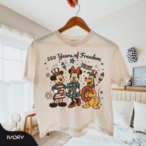 Może przedstawiać: Kremowa koszulka z Myszką Miki, Myszka Minnie i Pluto w patriotycznych strojach. Projekt zawiera napis "250 Years of Freedom" oraz świąteczne fajerwerki i gwiazdy. Koszulka wisi na drewnianym wieszaku.