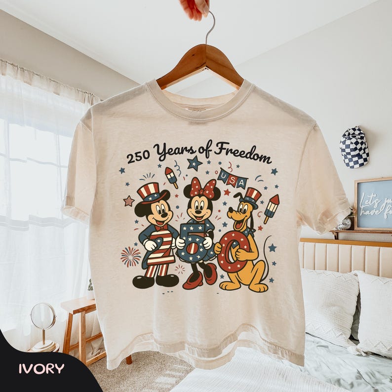 Może przedstawiać: Kremowa koszulka z Myszką Miki, Myszka Minnie i Pluto w patriotycznych strojach. Projekt zawiera napis "250 Years of Freedom" oraz świąteczne fajerwerki i gwiazdy. Koszulka wisi na drewnianym wieszaku.