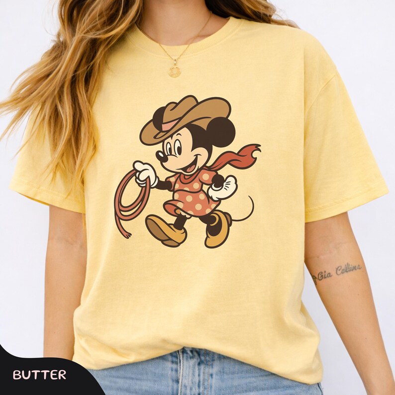 May include: Yellow t-shirt featuring a cartoon character in a cowboy hat, red scarf, and polka-dot dress, holding a rope. The word "BUTTER" is printed at the bottom left corner of the shirt. The shirt is made of a soft material.