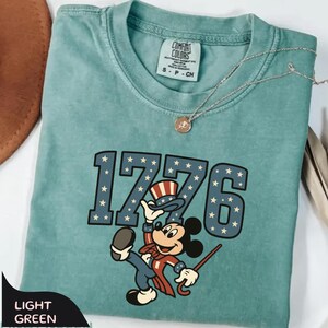 May include: Light green t-shirt with the number "1776" in blue with white stars, and a cartoon character in patriotic attire. The shirt has a Comfort Colors tag and a small gold necklace.