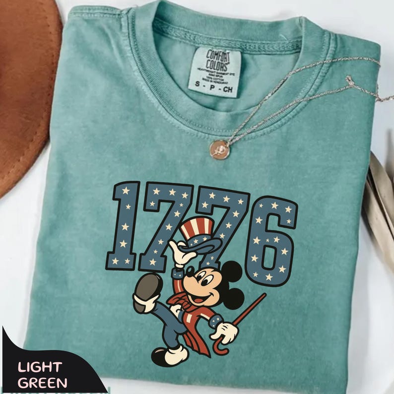 May include: Light green t-shirt with the number "1776" in blue with white stars, and a cartoon character in patriotic attire. The shirt has a Comfort Colors tag and a small gold necklace.