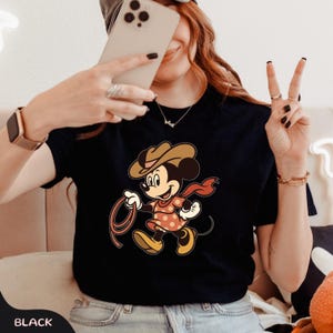 May include: Black t-shirt featuring a cartoon Minnie Mouse dressed as a cowgirl, wearing a brown hat, red scarf, and a polka-dot dress. Minnie is holding a lasso. The word "BLACK" is printed at the bottom of the shirt.