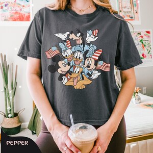 May include: Dark grey t-shirt featuring a graphic of Mickey Mouse, Minnie Mouse, Donald Duck, Daisy Duck, Goofy, and Pluto. The characters are wearing patriotic attire and holding American flags. The shirt is a comfortable, casual style.