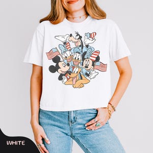 May include: White t-shirt featuring a graphic of Mickey Mouse, Minnie Mouse, Donald Duck, Daisy Duck, Goofy, and Pluto holding American flags. The characters are in patriotic colors. The word "WHITE" is at the bottom left.