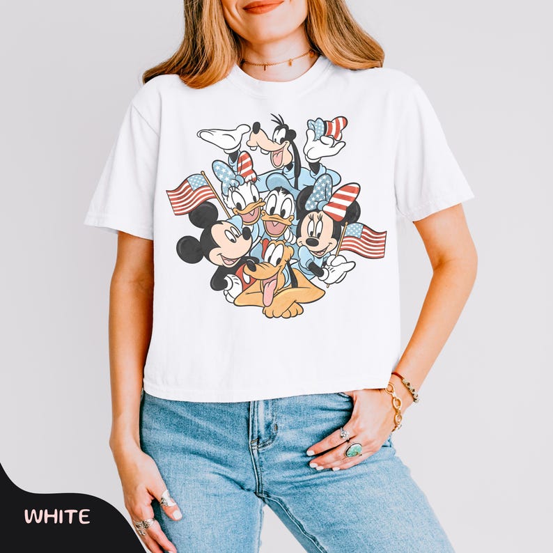 May include: White t-shirt featuring a graphic of Mickey Mouse, Minnie Mouse, Donald Duck, Daisy Duck, Goofy, and Pluto holding American flags. The characters are in patriotic colors. The word "WHITE" is at the bottom left.