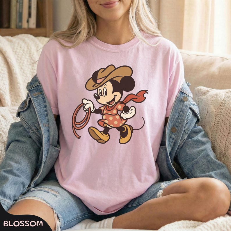 May include: A light pink t-shirt featuring a cartoon Minnie Mouse dressed as a cowgirl. She wears a brown hat, a red scarf, and a polka-dot dress, holding a lasso. The shirt is worn by a person.