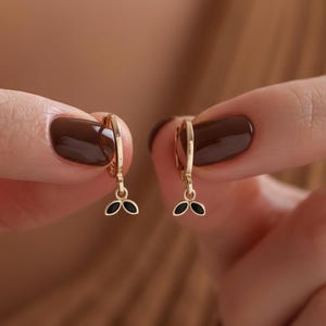 Könnte beinhalten: Ein Paar goldfarbene Creolen, jeweils mit einem kleinen, schwarzen, blattförmigen Anhänger. Die Ohrringe werden vor einem unscharfen Hintergrund gehalten, der ihr zartes Design und ihre elegante Schlichtheit zeigt.