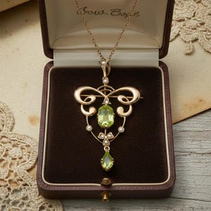 Pode incluir: Um colar com pingente dourado com uma pedra preciosa peridoto e pequenas pedras brancas, exibido em uma caixa de joias marrom. O pingente tem um design ornamentado e simétrico com um peridoto em forma de gota na parte inferior.