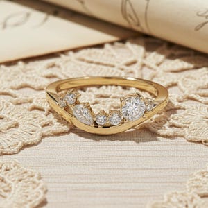 May include: A gold ring with a curved band, set with multiple clear gemstones of varying sizes. The ring's design features a cluster of stones, including round and marquise cuts. The ring is displayed on a lace surface.