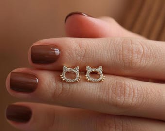 May include: A pair of gold-colored cat-shaped stud earrings. Each earring features a cat face design with small, clear gemstones outlining the face. The earrings are small and delicate, suitable for everyday wear.