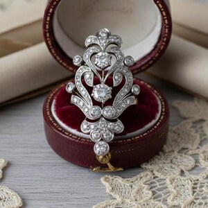 May include: An ornate, silver-toned brooch with numerous clear gemstones, displayed open in a burgundy jewelry box with a red velvet interior. The brooch is surrounded by antique scrolls and lace.