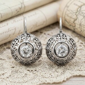 May include: A pair of silver-toned filigree earrings with a central, round, clear gemstone. The earrings have a lever back closure and are displayed on a lace surface, with antique paper in the background.