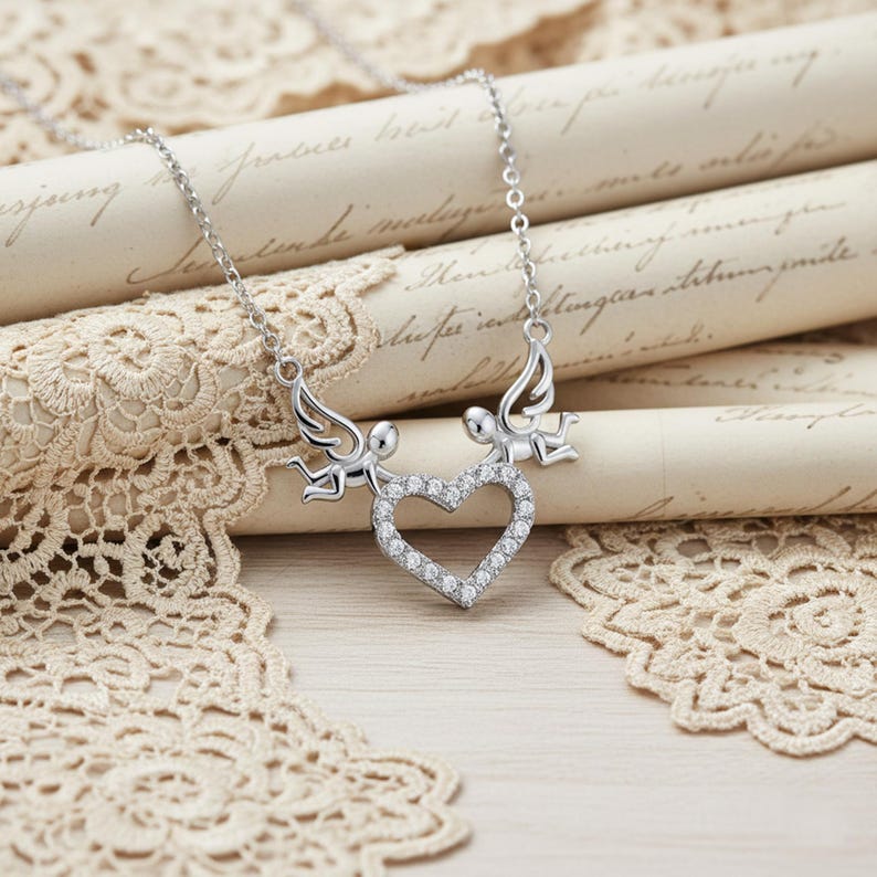May include: A silver necklace featuring two angel figures holding a heart-shaped pendant. The heart is encrusted with small, clear gemstones. The necklace is displayed on a backdrop of vintage paper and lace.