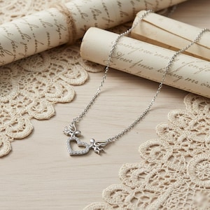 May include: A silver necklace featuring a heart pendant with a halo of small, clear stones. Two small angel figures flank the heart, and the necklace chain is delicate. The necklace rests on a light-colored surface with lace and rolled paper scrolls in the background.