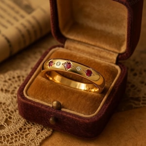 May include: A gold ring with a series of red gemstones and small clear stones set in a band. The ring is displayed in a burgundy velvet jewelry box with a gold interior. The ring's design includes star-shaped settings around the red stones.