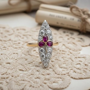 May include: A gold ring with an elongated, oval setting. The setting is studded with small, clear gemstones and features a three-leaf clover design with three red gemstones. The ring rests on a cream-coloured lace doily.