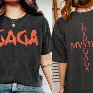 May include: Dark gray t-shirt with the word "GAGA" in distressed orange lettering on the front. The back of the shirt features a cross design with the words "LVD MAYHEM GAGA" in orange.