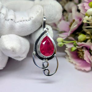 Puede incluir: Un colgante de plata con un gran rubí facetado en forma de lágrima. El rubí es de color rojo vibrante y está engastado en un marco de plata decorativo con un lazo en la parte superior para una cadena. El colgante tiene un diseño estilizado y abierto con un detalle en espiral.