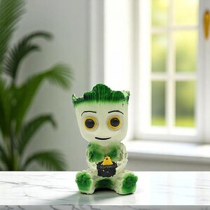 May include: A small, seated figurine of a cartoon character with a white body, green accents, and large yellow eyes. The figurine is holding a black object with a yellow bird on top. The figurine is on a white surface.