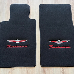May include: Two black car floor mats with the Thunderbird logo embroidered in red and white. The mats are rectangular with a contoured driver-side mat. The Thunderbird logo is centered on each mat.