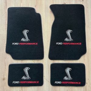 May include: Four black car floor mats with a white cobra logo and the words "FORD PERFORMANCE" in red. The mats are designed to protect a vehicle's floor. The set includes two front and two rear mats.