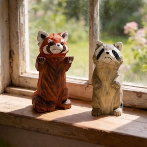 May include: Two wooden animal figurines, a red panda and a raccoon, stand on a windowsill. The red panda is reddish-brown with a white face and ears. The raccoon is gray and white. Both are carved with detailed features.