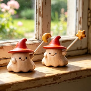 Pode incluir: Duas figuras de fantasmas de madeira com corpos de madeira clara lisa e pequenos rostos sorridentes. Cada fantasma usa um chapéu vermelho pontudo. Um segura uma varinha com uma lua crescente amarela, o outro uma varinha com uma estrela amarela.