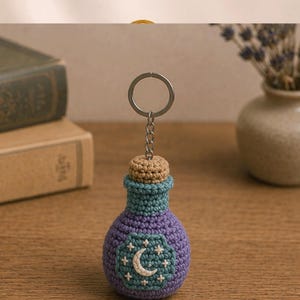May include: A handmade crochet keychain shaped like a potion bottle. The bottle is purple with a teal neck and a tan cork. A crescent moon and star design is on the front. The keychain is attached to a silver-colored ring.