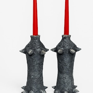 May include: Two dark gray, textured candle holders with red candles. The holders have a unique design with protruding, spiky elements. The candles are tall and slender, providing a striking contrast to the holders.