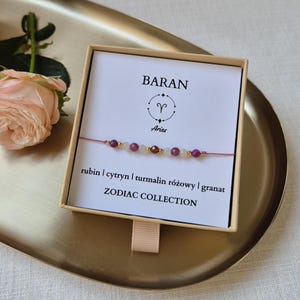 Può includere: Braccialetto della collezione Zodiaco Ariete con perline di rubino, citrino e tormalina rosa. Il braccialetto è presentato in una confezione regalo con il simbolo dell'Ariete e testo. Una rosa rosa è sullo sfondo.