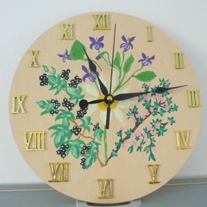 Peut inclure: Une horloge murale ronde avec un cadran beige clair, des chiffres romains dorés et des aiguilles noires. Le cadran est décoré d'un motif floral vert, violet et blanc. L'horloge mesure environ 30 cm de diamètre.
