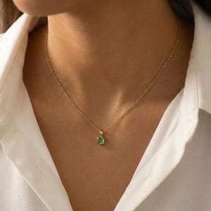 May include: A gold-colored necklace with a small, square-cut, light green gemstone pendant. The necklace is delicate and appears to be made of thin chain links. The pendant is centered on the chest, against a white shirt.