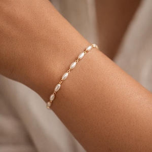 May include: A delicate gold bracelet featuring a series of oval-shaped pearls. The pearls are strung together with fine gold links, creating a simple yet elegant design. The bracelet is shown on a person's arm, suggesting it is a piece of jewelry.
