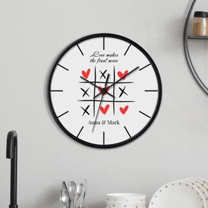 May include: A round wall clock with a black frame and white face. The clock features a tic-tac-toe design with red hearts and black X's. The text "Love makes the final move" and names "Anna & Mark" are printed on the clock face.