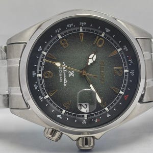 May include: Silver-toned wristwatch with a dark green dial featuring gold-colored numerals and hands. The watch has a date display at the 3 o'clock position and a stainless steel band. The watch face includes the text "Automatic 20BAR".