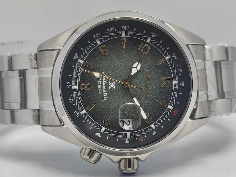 May include: Silver-toned wristwatch with a dark green dial featuring gold-colored numerals and hands. The watch has a date display at the 3 o'clock position and a stainless steel band. The watch face includes the text "Automatic 20BAR".
