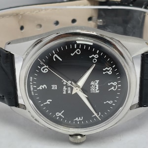 May include: A wristwatch with a black dial and silver-tone case. The watch features Arabic numerals and hands, with a black leather band. The watch face has text in a foreign language. The watch is a classic design.