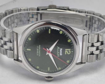 May include: Close-up of a silver wristwatch with a dark green face. The watch has a silver band and features the text "hmt Pilot" and "Para Shock 17 Jewels". The watch hands are white with a red second hand.