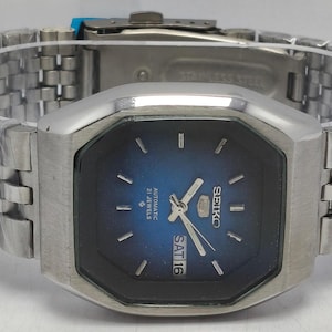May include: A Seiko 5 automatic watch with a silver-tone stainless steel link bracelet and a blue gradient dial. The watch features a hexagonal case, silver-tone hands and hour markers, and a day-date window at the three o'clock position.