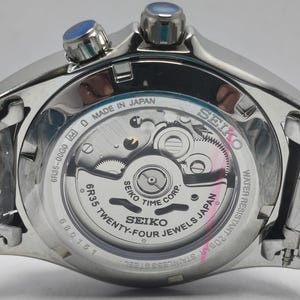 May include: Close-up of a silver Seiko watch with a transparent back, revealing the intricate inner workings. The watch features blue accents on the crown and is labeled "Made in Japan." The watch is water resistant and made of stainless steel.