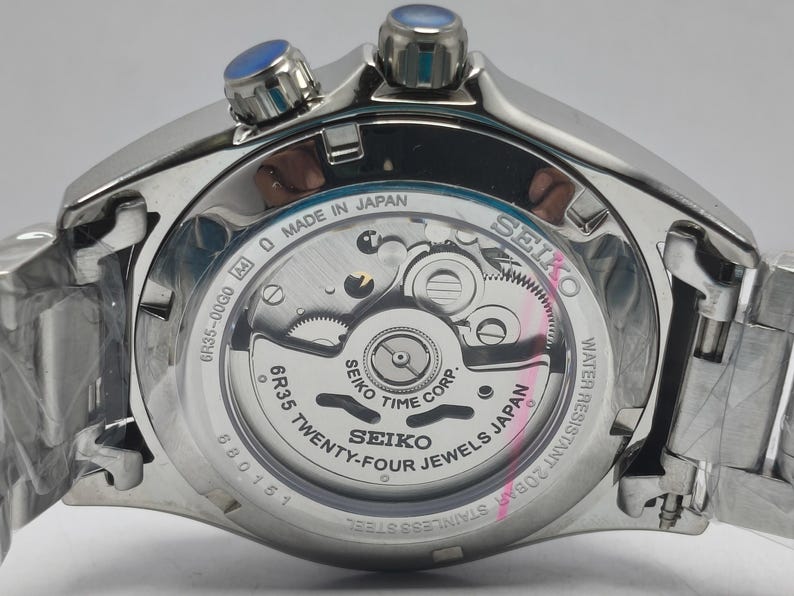 May include: Close-up of a silver Seiko watch with a transparent back, revealing the intricate inner workings. The watch features blue accents on the crown and is labeled "Made in Japan." The watch is water resistant and made of stainless steel.