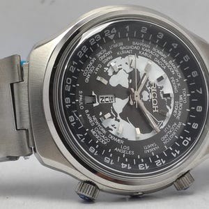 May include: A silver-toned wristwatch with a black face featuring a world map design and a 24-hour time display. The watch has a stainless steel band and a Ricoh logo. The watch also displays the time zones of major cities.