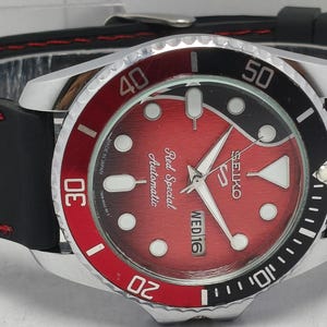 Op de afbeelding: Een Seiko Red Special Automatic horloge met een zwarte rubberen band en rode stiksels. De wijzerplaat is rood met witte markeringen en wijzers. De lunette is zwart en rood, met witte cijfers.