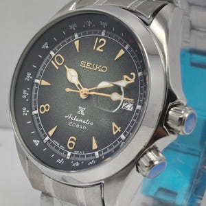 May include: A stainless steel Seiko automatic wristwatch with a black dial. The watch features gold-colored hands and hour markers, a date display, and a rotating bezel. The watch band is made of stainless steel.