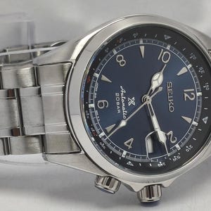 May include: A silver-toned stainless steel Seiko automatic watch with a deep blue dial. The watch features a metallic link bracelet, a rotating compass bezel, luminous hands, and a date window at the three o'clock position with a cyclops lens.