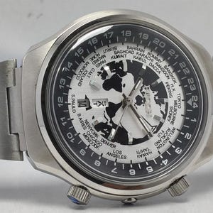 May include: A silver-toned wristwatch with a black and white world map design on the face. The watch features a rotating bezel with city names and a date display. The watch band is also silver-toned.